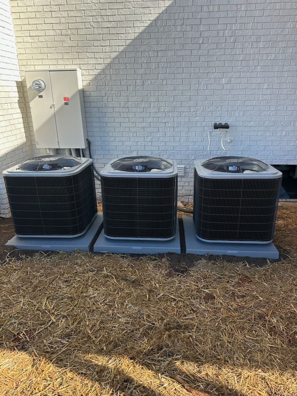 Three identical black air conditioning units sit on gray pads against a painted brick wall.