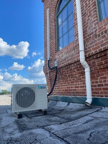 A Daikin outdoor AC unit sits on a flat rooftop next to a red brick building with a large window and white drainpipe.
