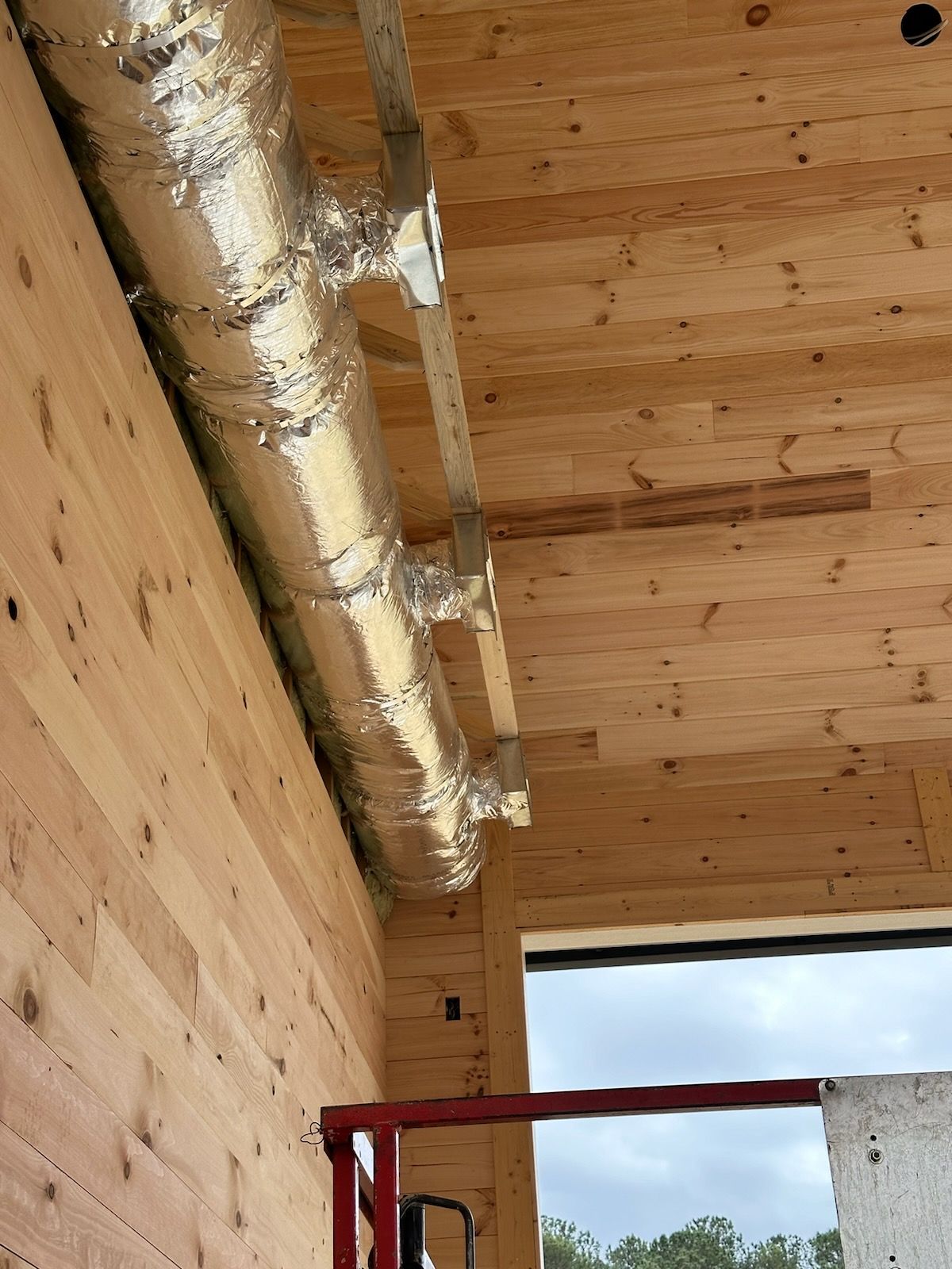 A silver insulated HVAC duct runs along the corner where wooden wall panels meet a matching timber ceiling.