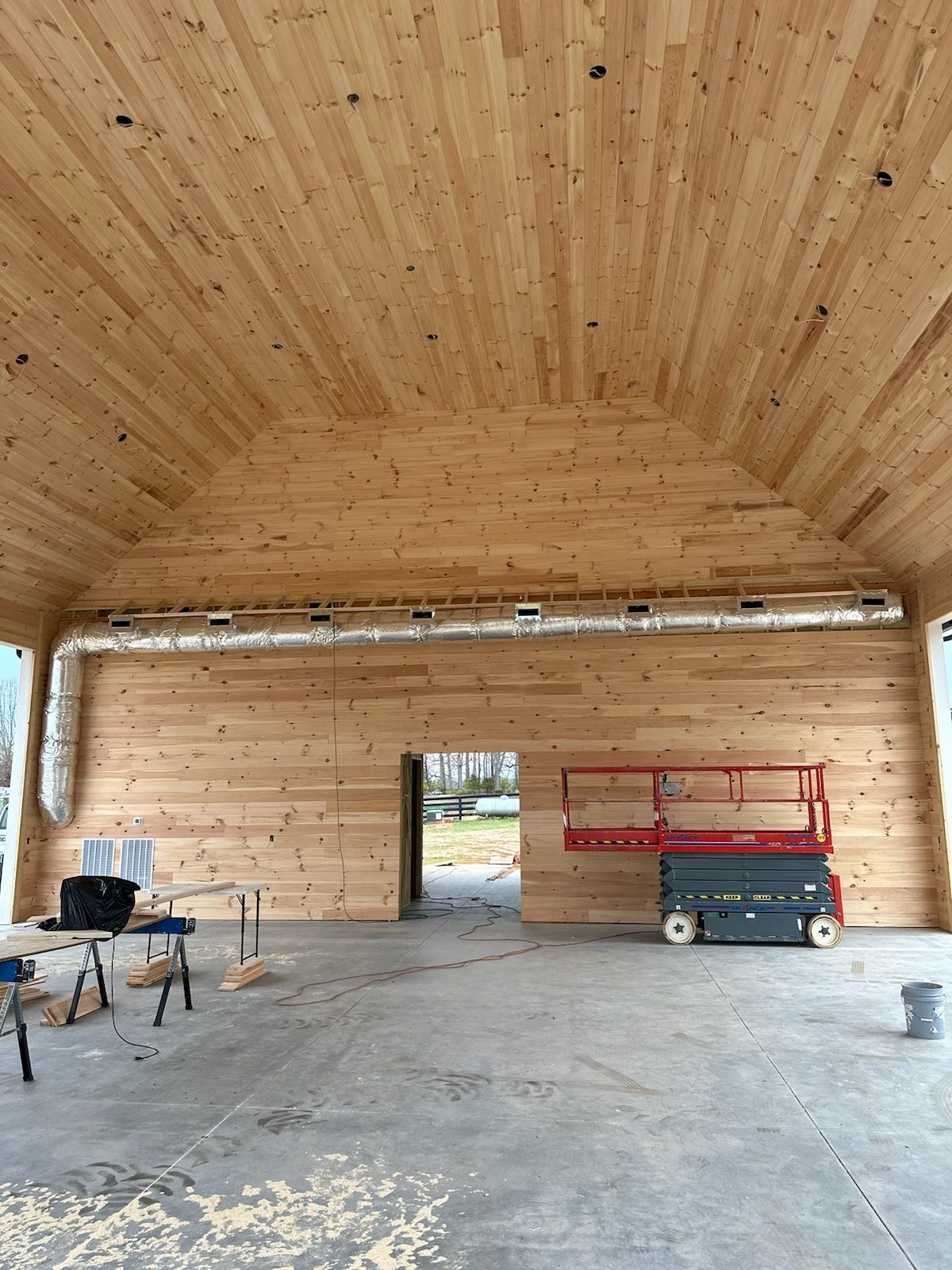 An interior view of a large, unfinished wooden building with vaulted ceilings, exposed metal ductwork, and a scissor lift.