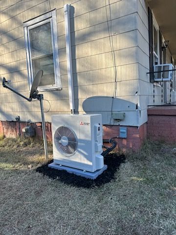 A Mitsubishi mini-split heat pump outdoor unit sits on a pad against the exterior wall of a house near a satellite dish.