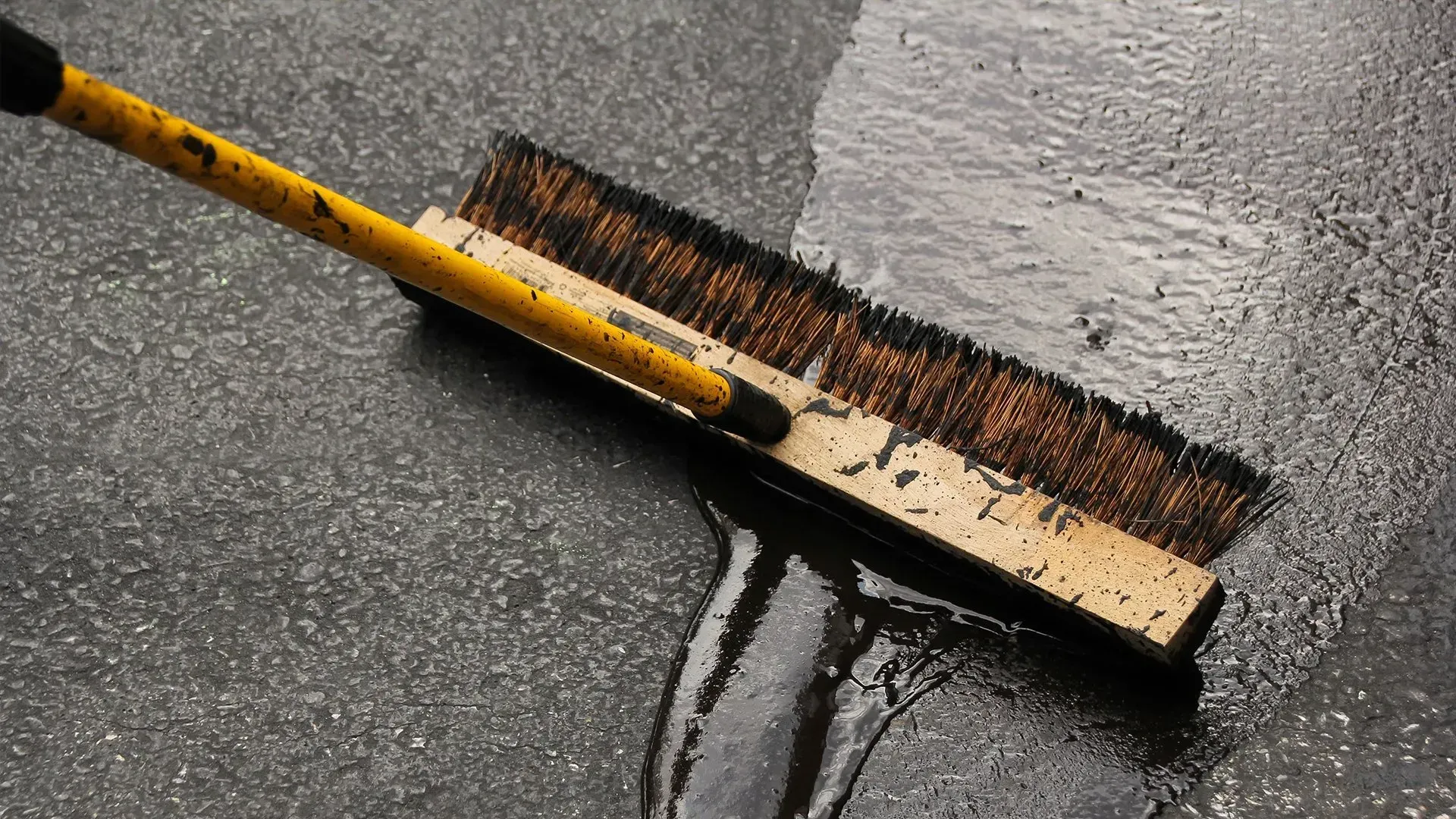 Broom sweeping water and grime across wet pavement, with a yellow handle and dark bristles.