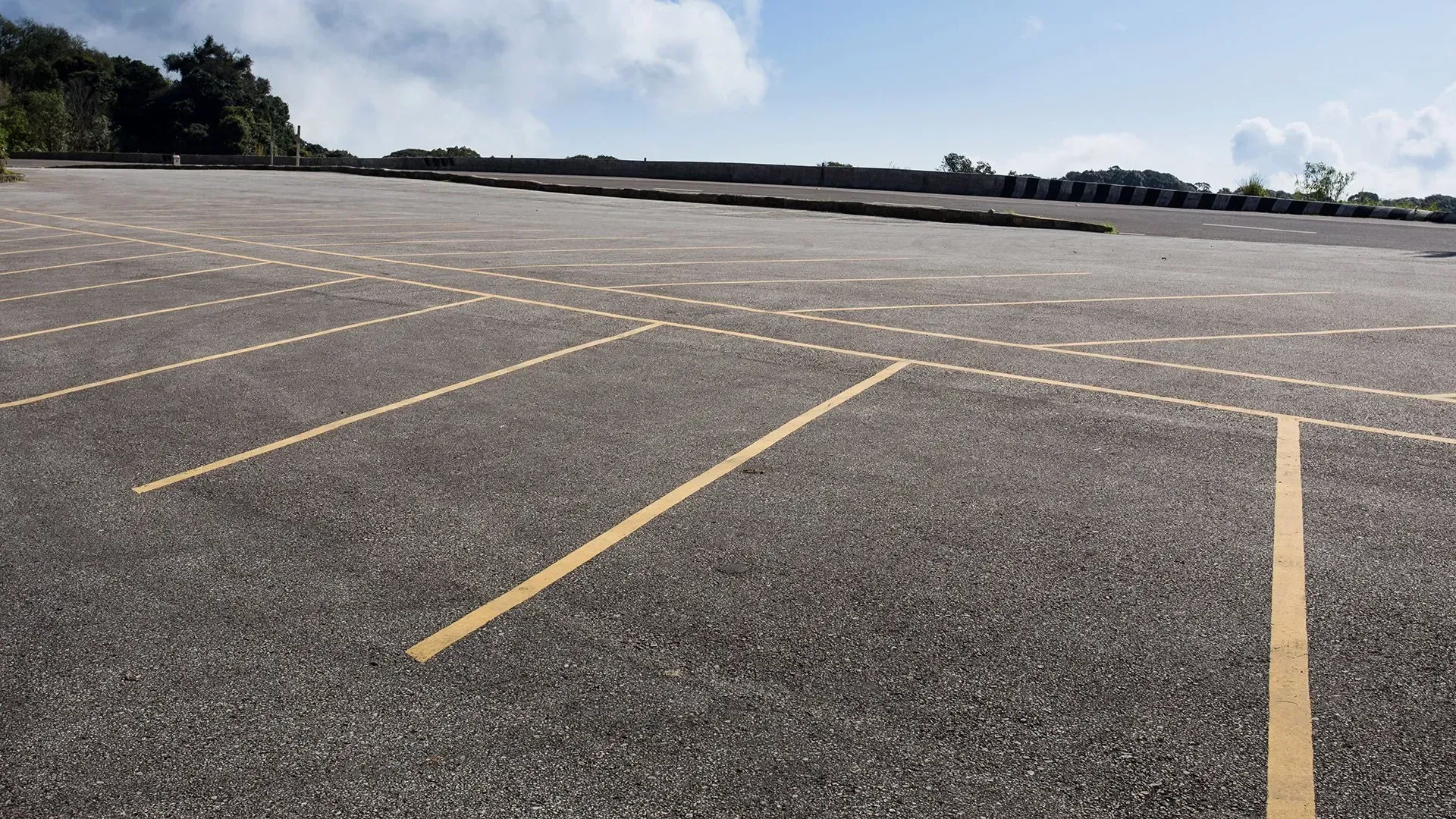 Empty asphalt parking lot with yellow parking lines under a bright, cloudy sky.