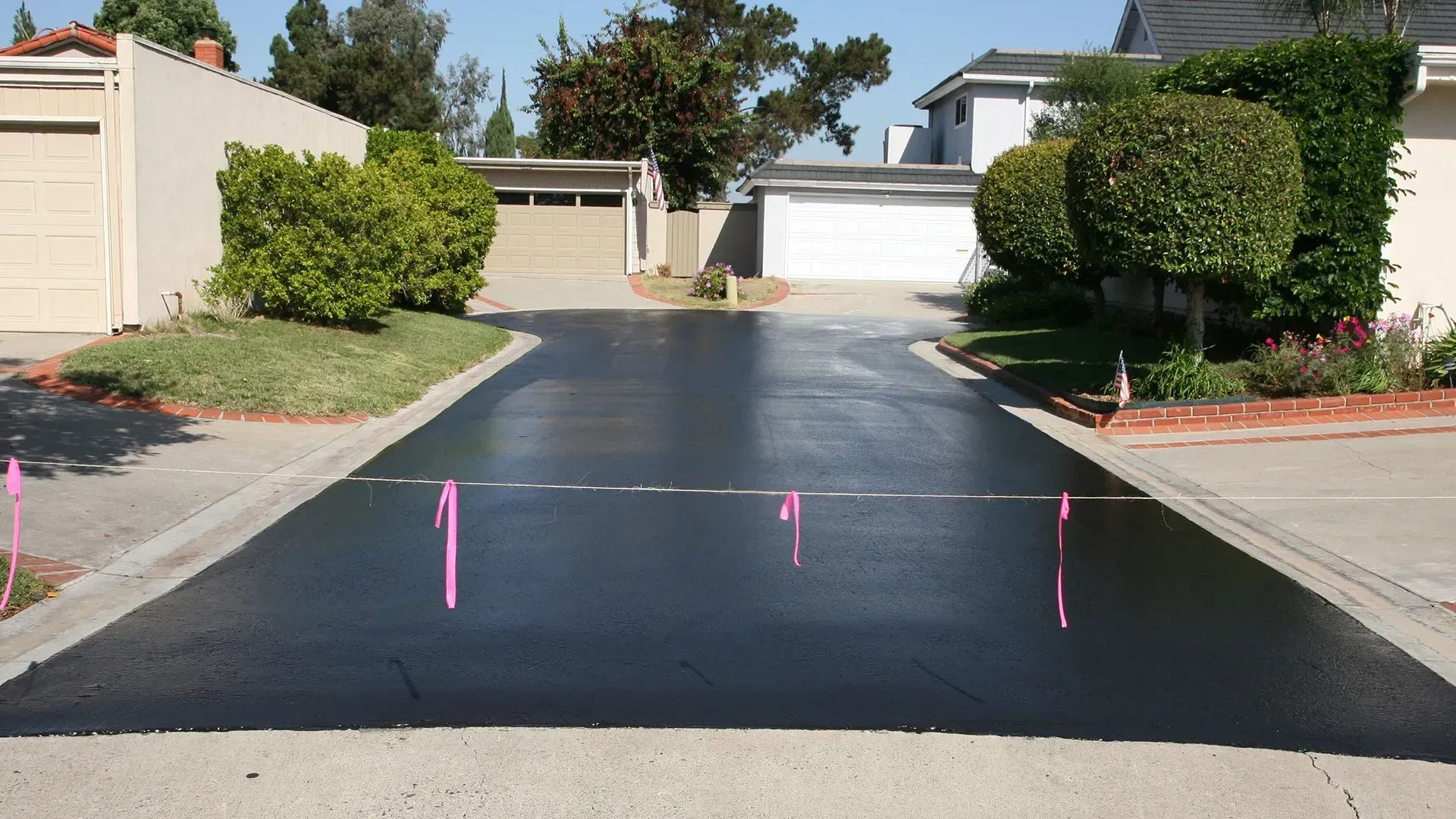 Residential driveway freshly sealed black, with pink markers, between houses and bushes