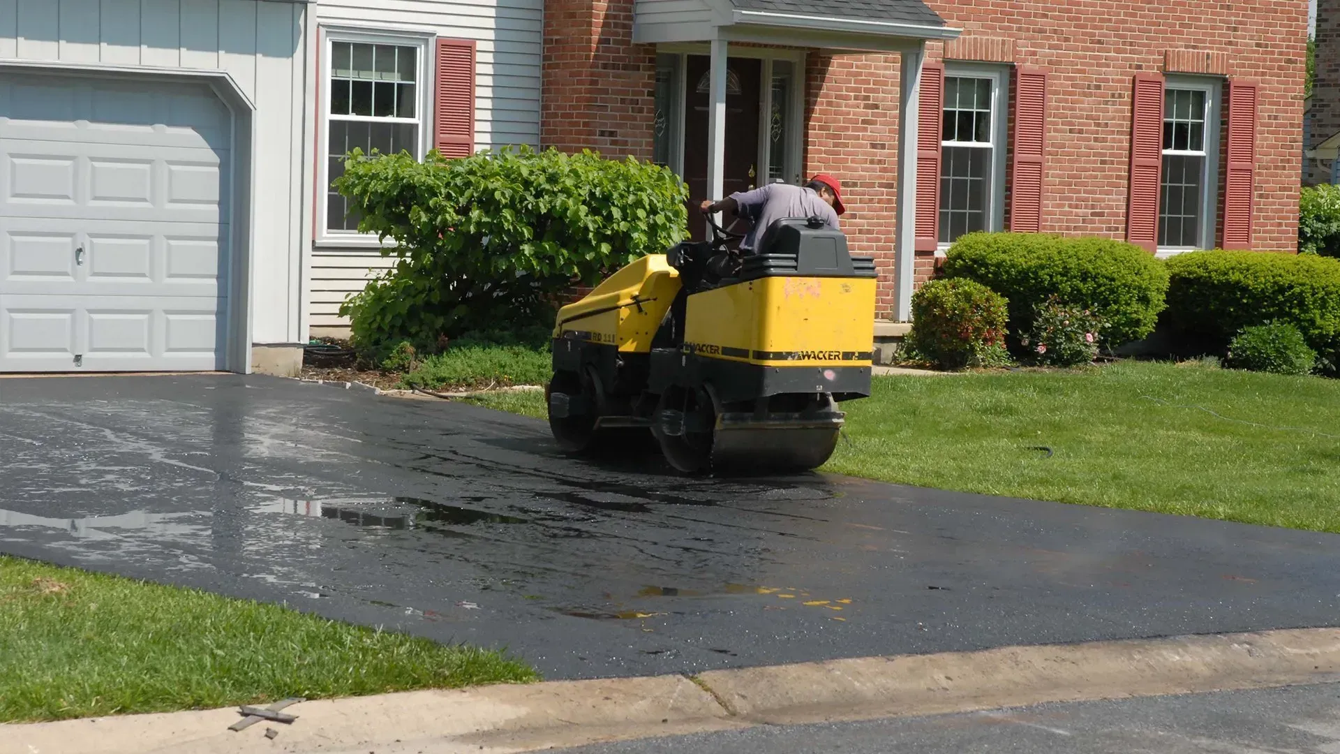 Yellow robotic lawn mower on a wet driveway beside a suburban house and garage