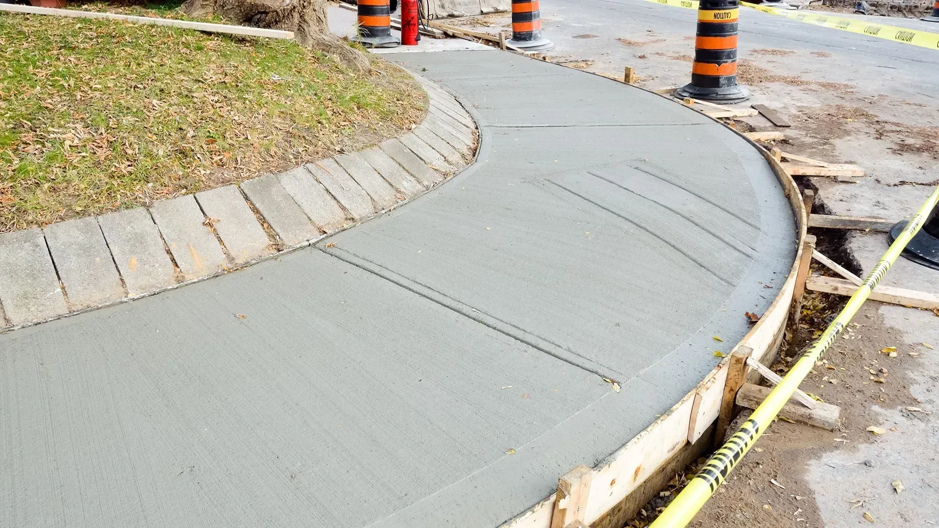 New concrete sidewalk curve beside grass and roadwork cones, with caution tape along the edge