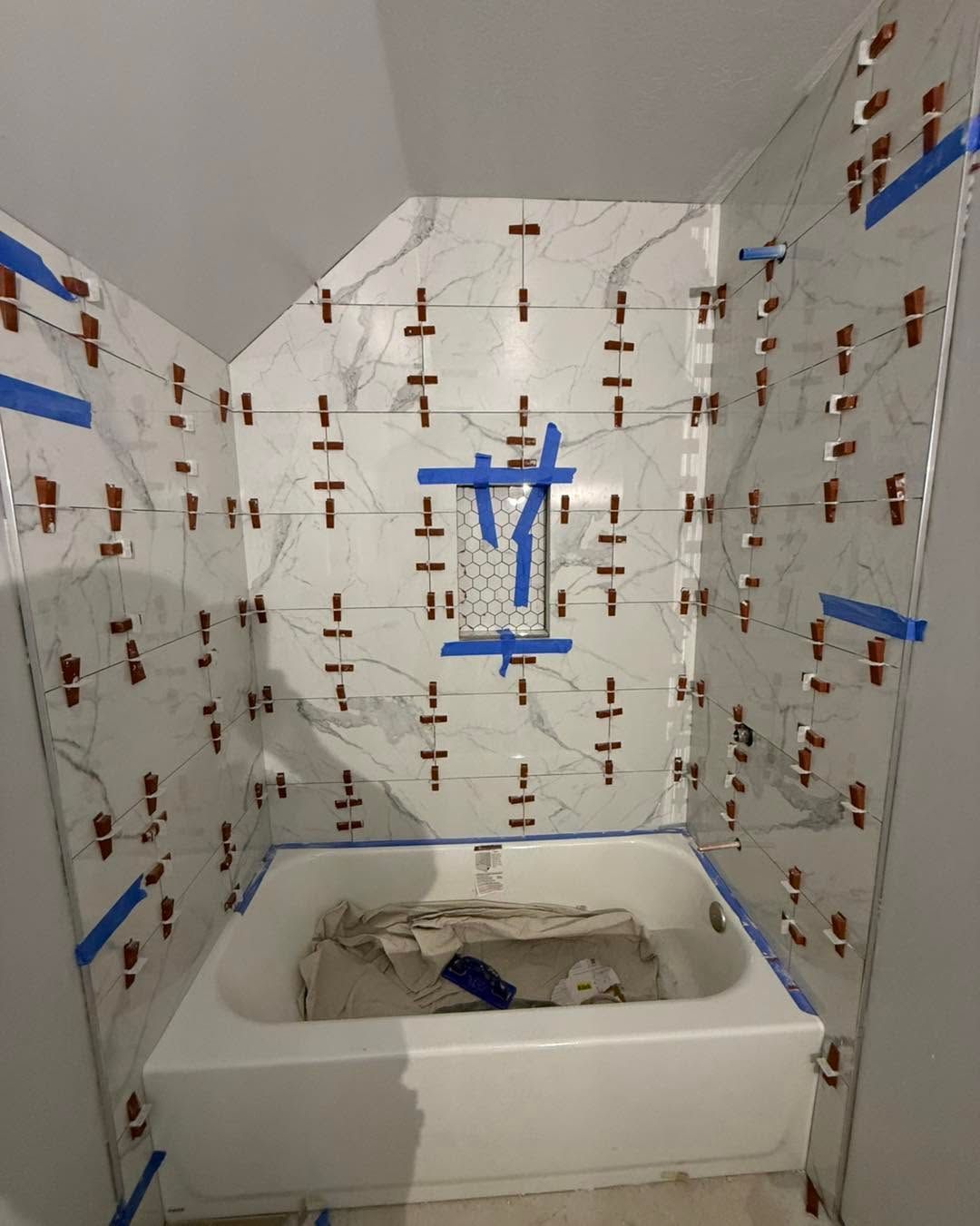 Partially tiled bathroom with a white bathtub under renovation, covered in protective plastic and blue tape.