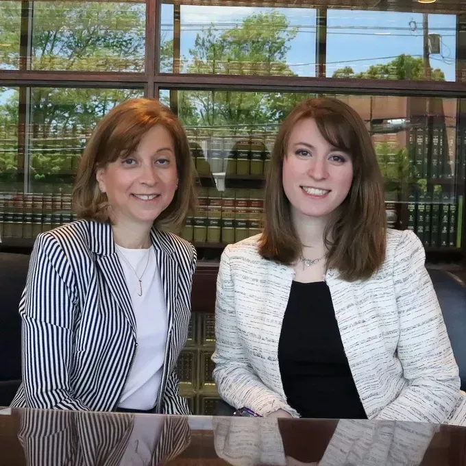 Two women in business attire seated at a table, smiling at the camera. Office setting with window.