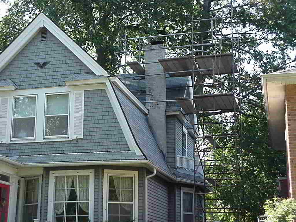 Gray house with exterior scaffolding and a brick chimney amid trees
