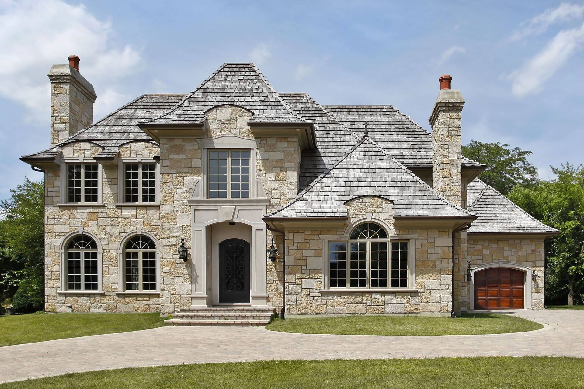 Large stone suburban house with a shingled roof, arched windows, and a two-car garage under a cloudy sky
