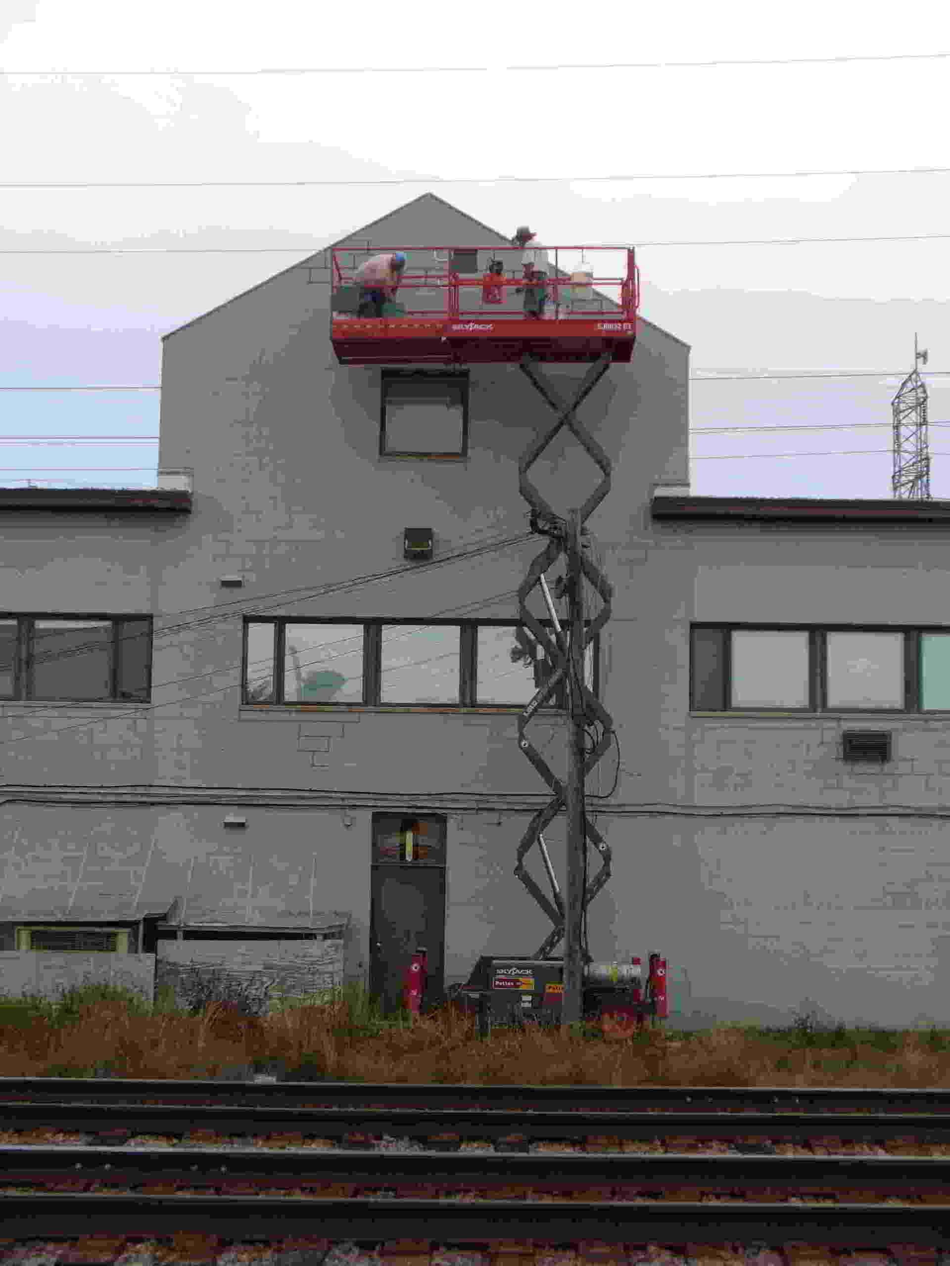 Gray building with a raised red scissor lift by railroad tracks