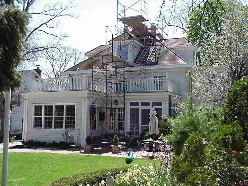 White house with porch and scaffolding in a sunny yard, surrounded by trees and green lawn