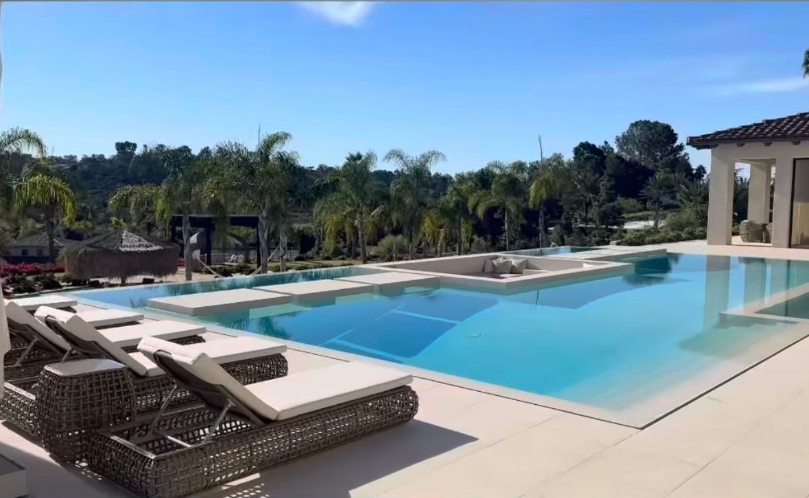An in ground pool in the daytime with a tree line in the distance and pool chairs at poolside.