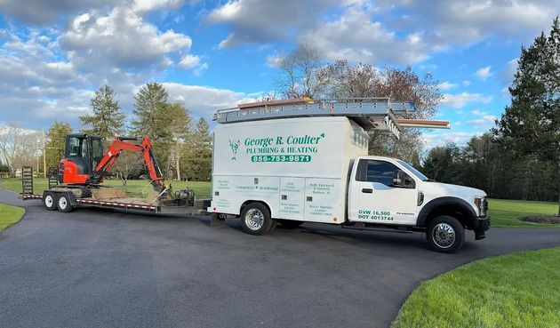 White landscaping truck towing an orange mini excavator on a flatbed trailer in a driveway under a cloudy sky.