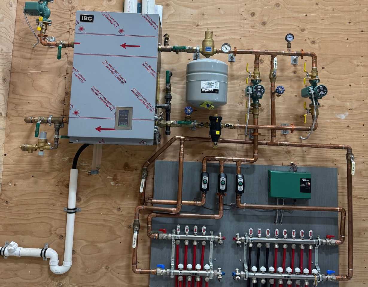 Wall-mounted plumbing and heating manifold with pipes, valves, tanks, and control boxes on a plywood board.