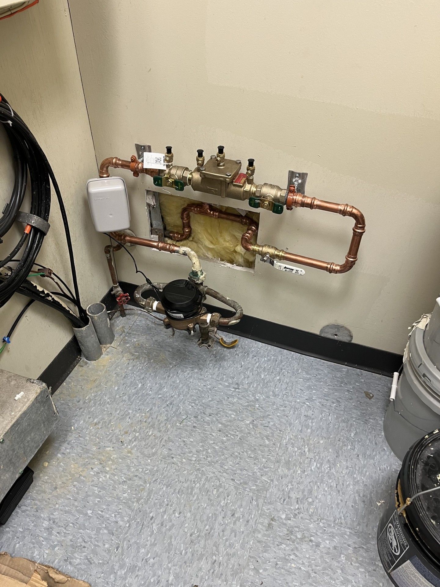 Mechanical plumbing and copper piping on a wall in a utility room, with gauges and floor equipment below.