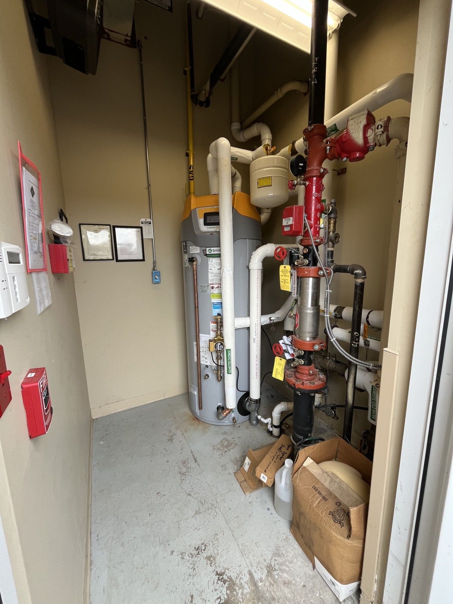 Utility room with pipes, valves, water heater, and stacked cardboard boxes in a narrow industrial space