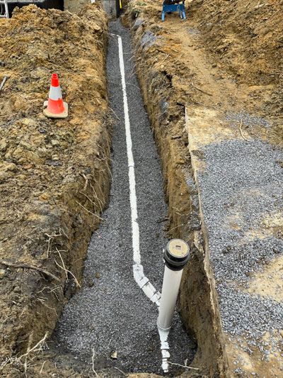 Trench with gravel-lined drainage pipe and white conduit beside a dirt excavation, marked by an orange cone.
