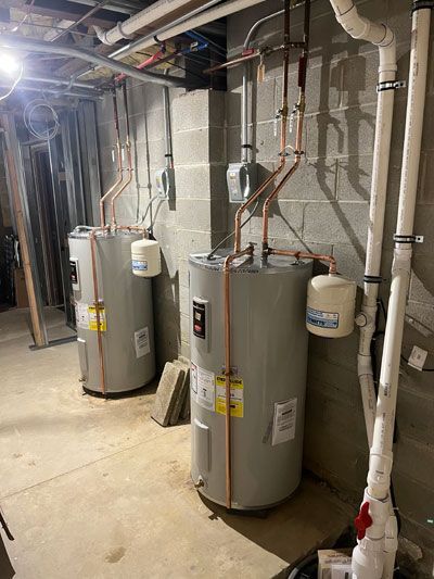 Water heater room with two gray tanks, exposed pipes, and utility equipment in a basement.
