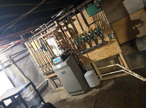 Utility room with boiler, gauges, and exposed pipes along a wall in a dim basement.
