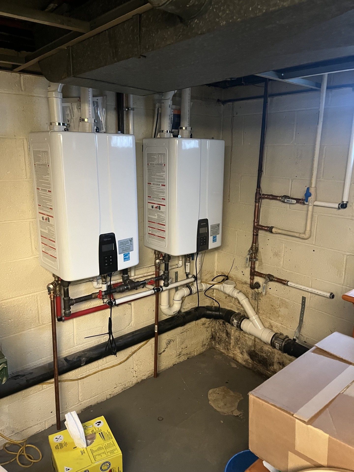 Two white wall-mounted water heaters in a basement utility room with exposed pipes and boxes.