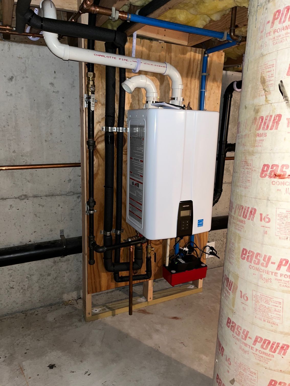 White tankless water heater mounted on framing in an unfinished basement, with exposed pipes and insulation.