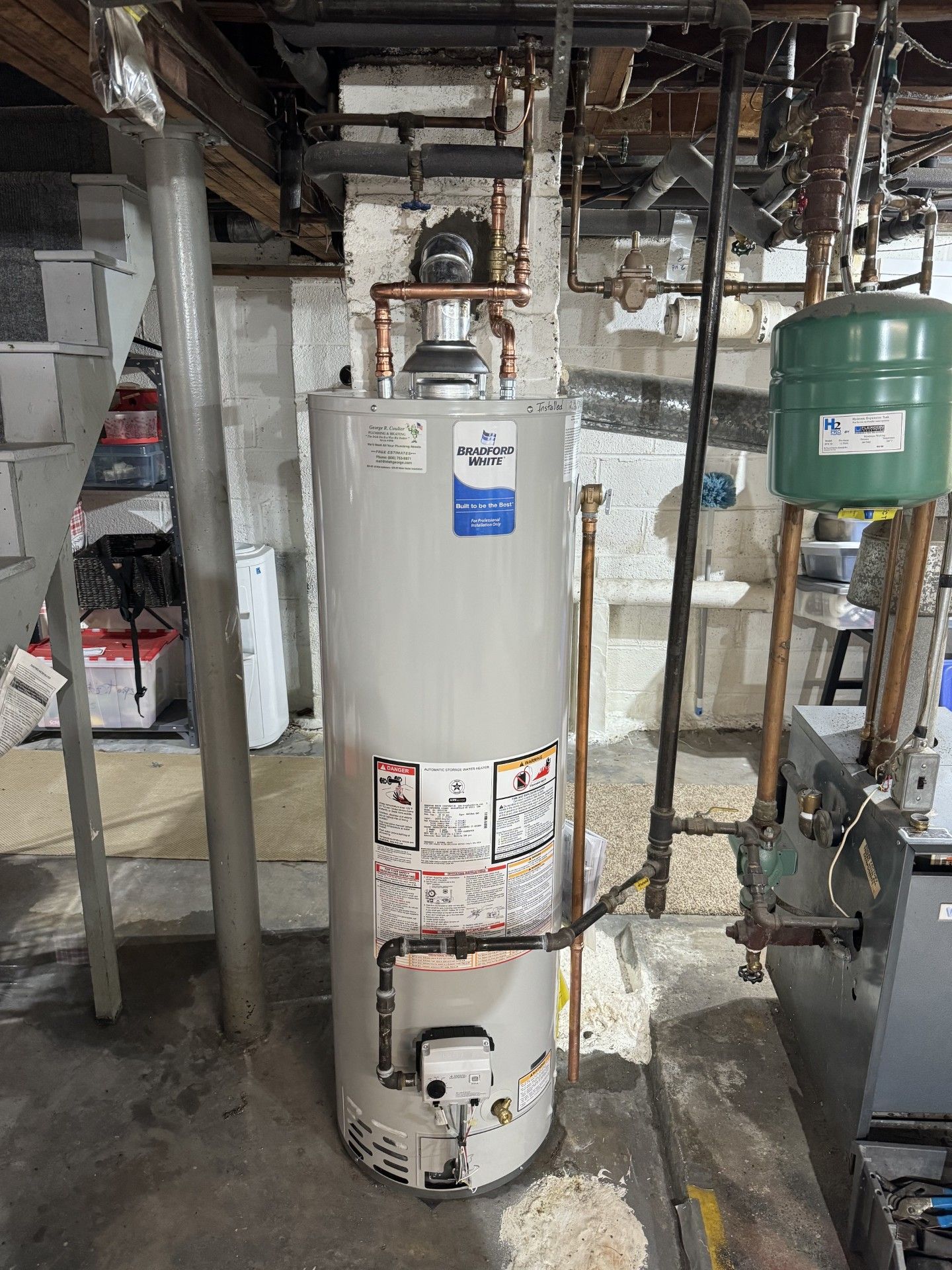 White water heater in a basement utility area with pipes, shelves, and a green tank nearby