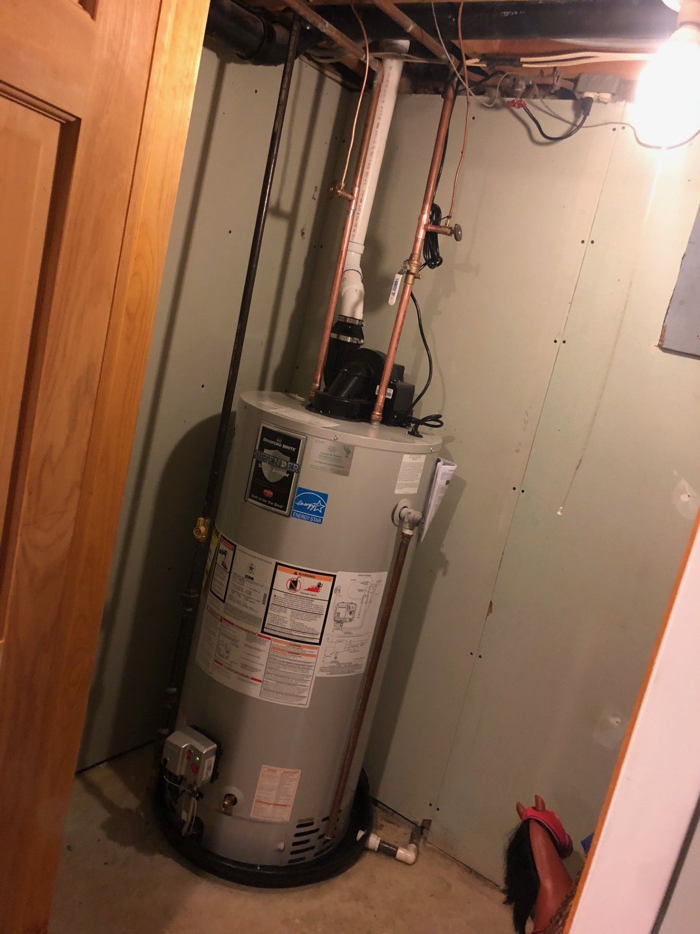 Gas water heater in a narrow utility closet with exposed pipes and a small red fire extinguisher.