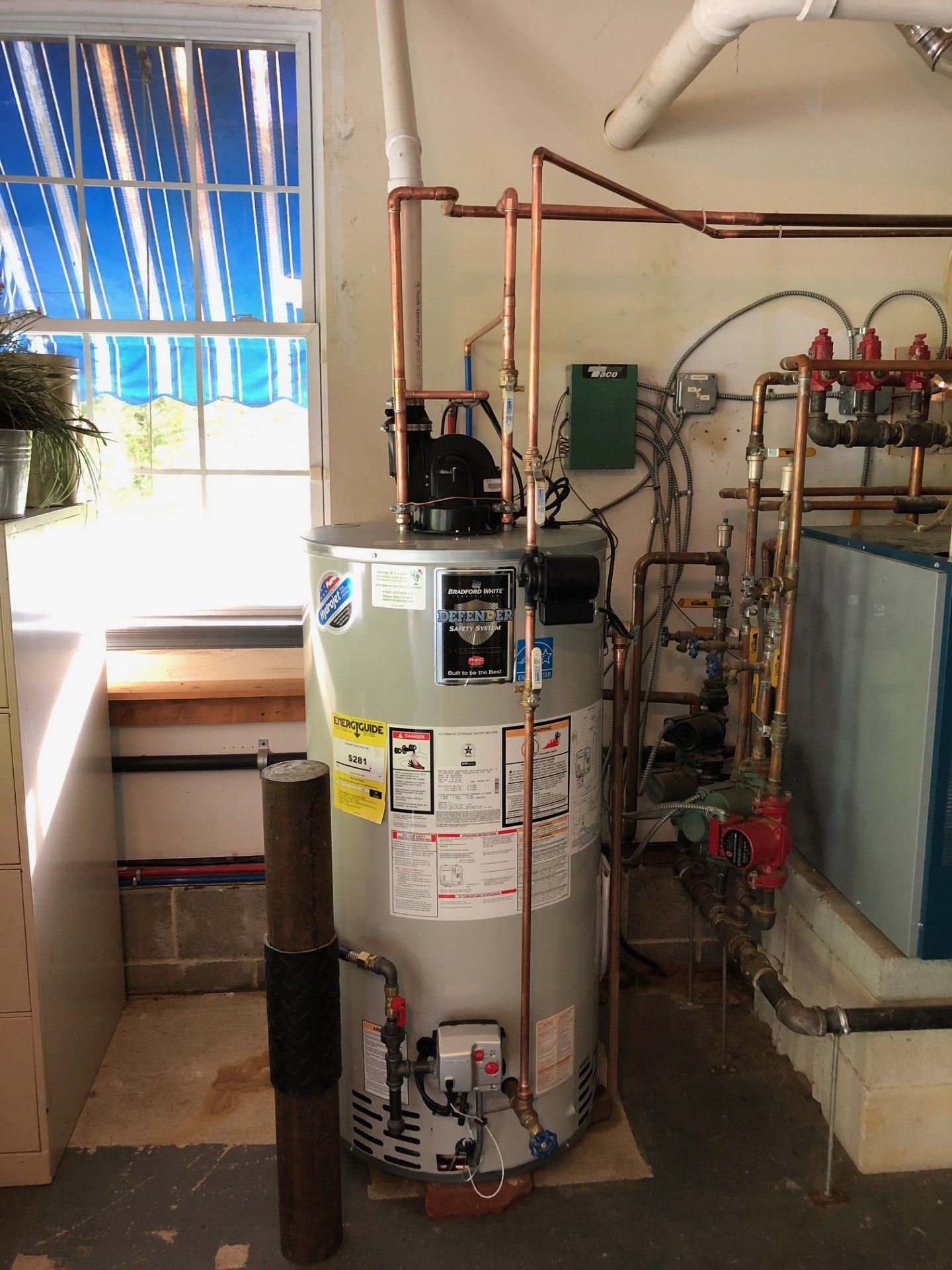 Water heater and plumbing equipment in a utility room, with pipes, gauges, and a window on the left.