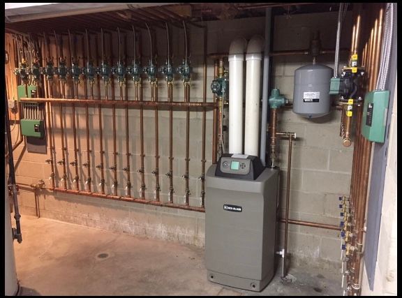 Basement furnace and boiler system with pipes, valves, and gray utility tanks along a cinder block wall