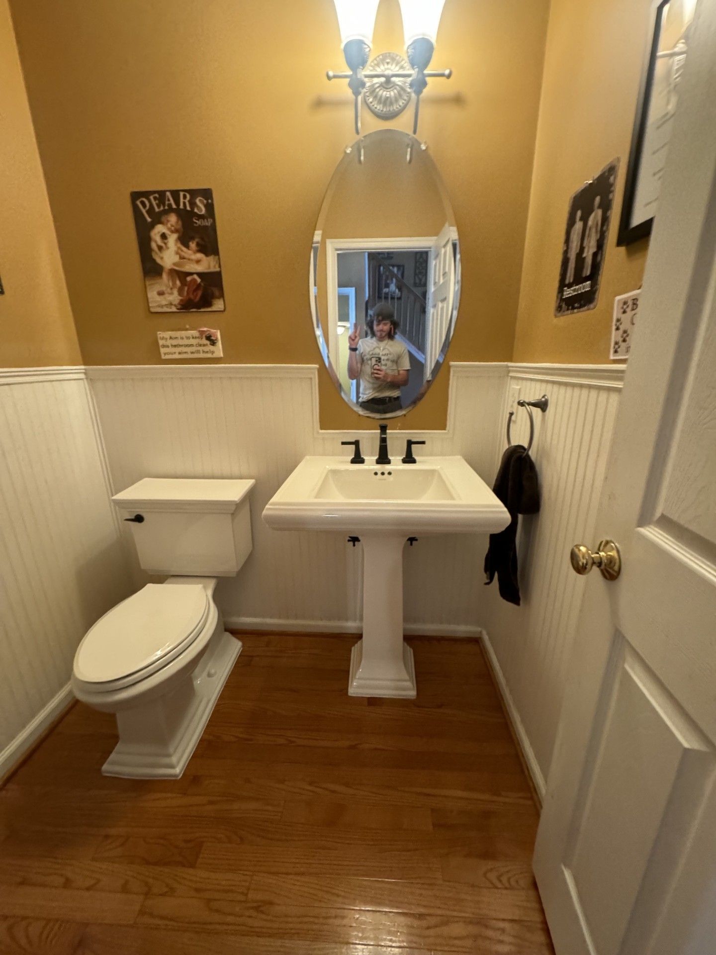 Small bathroom with pedestal sink, oval mirror, toilet, and wood floor under warm lighting.