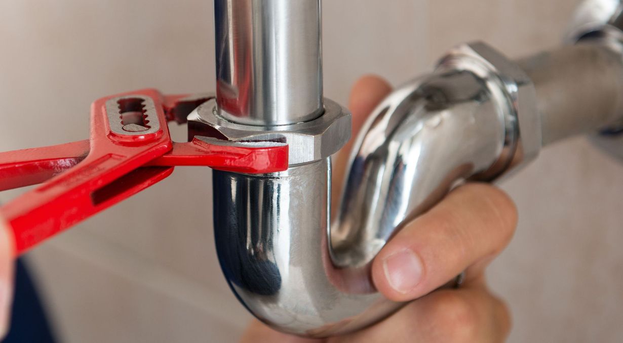 Red pipe wrench tightening a shiny chrome sink drain pipe under a sink.