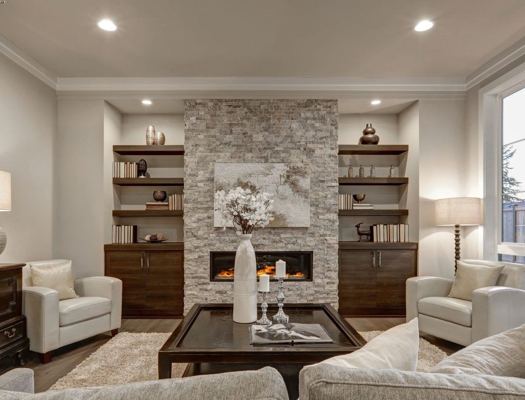 Elegant living room with stone fireplace, built-in shelves, and neutral armchairs around a coffee table.