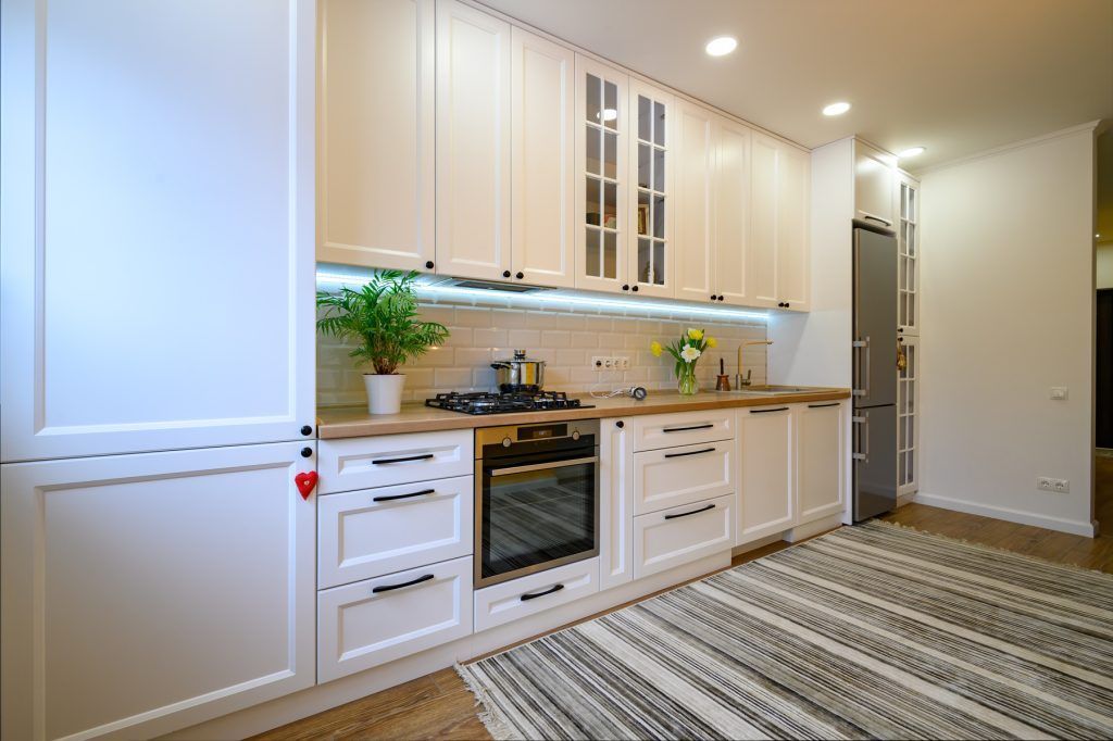Bright modern kitchen with white cabinets, built-in oven, and striped floor runner in a long hallway-style layout