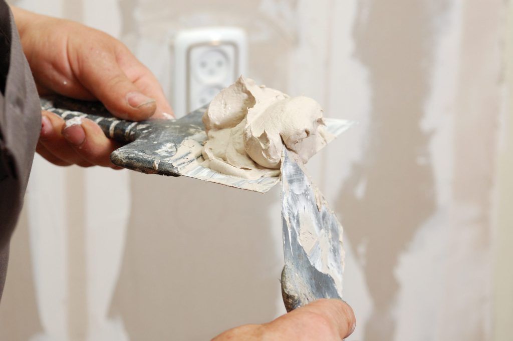 Hands using putty knives to spread drywall compound over a wall outlet area.
