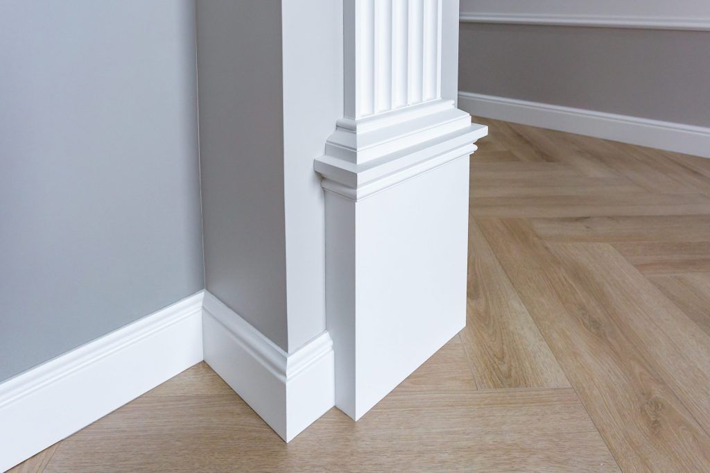 White column trim at a room corner with gray walls and light wood flooring.