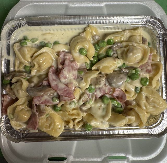 Tray of tortellini in creamy sauce with peas and pieces of ham in a foil container.
