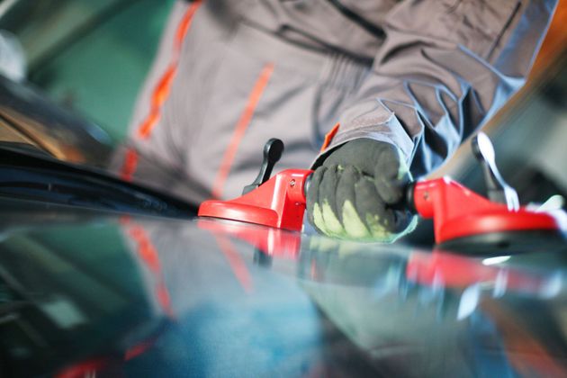 A worker wearing work gloves uses a red dual-suction tool to lift or position a car windshield.