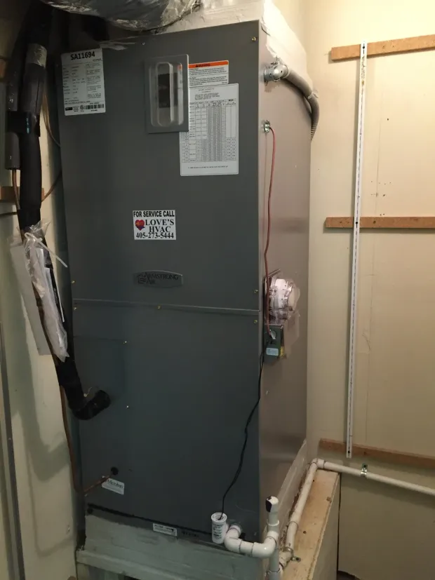 Gray furnace in a utility closet with exposed pipes and wiring beside a beige wall.