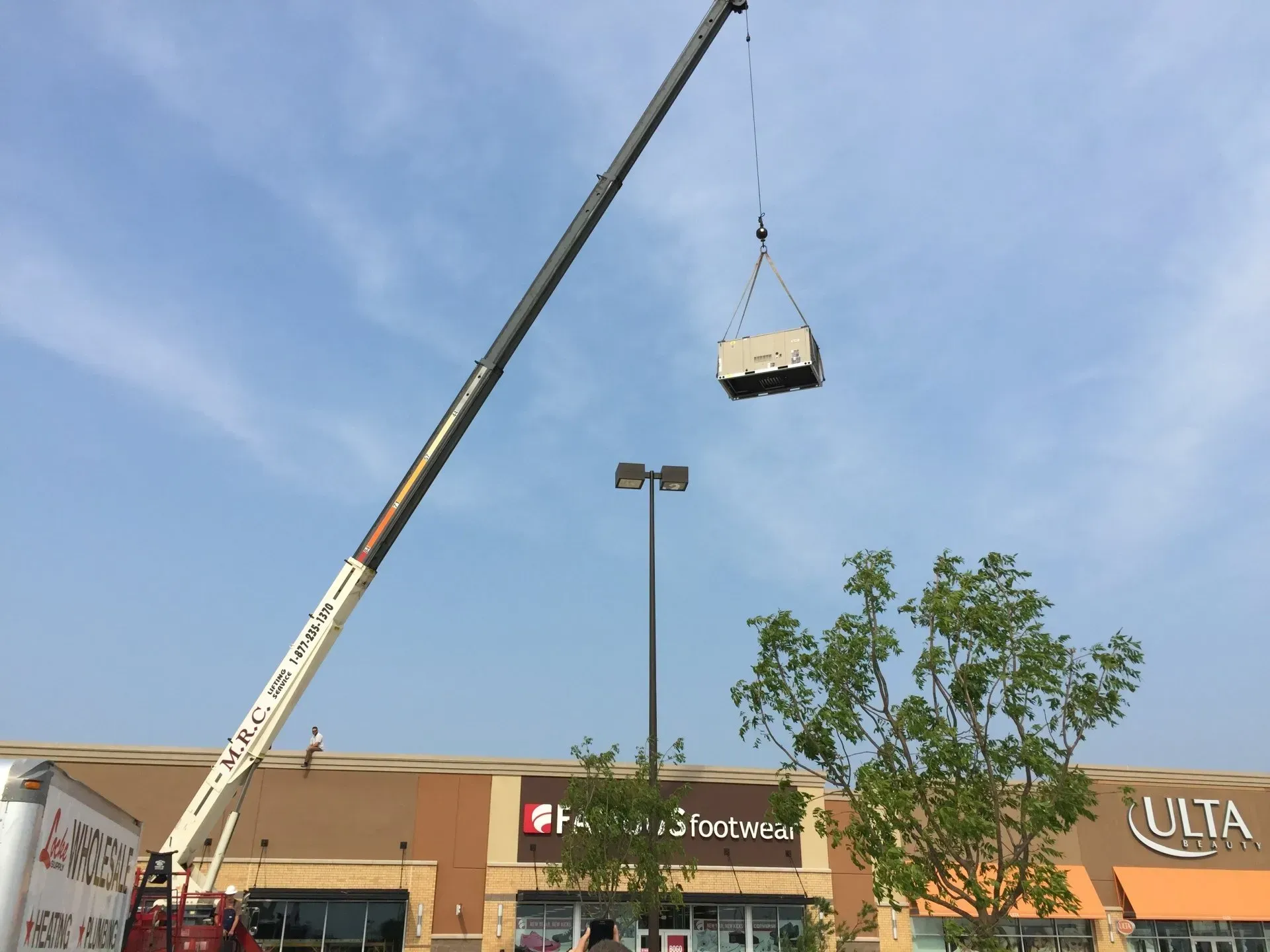 Crane lifting a large rectangular object above a shopping center parking lot under a blue sky