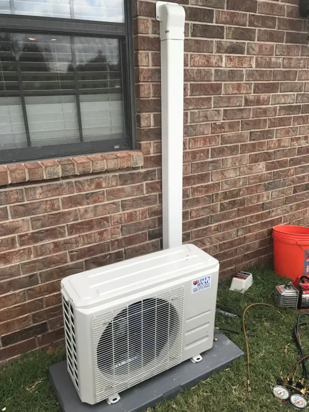 Outdoor air conditioner unit beside a brick house wall under a window, with a white drain pipe and red bucket nearby