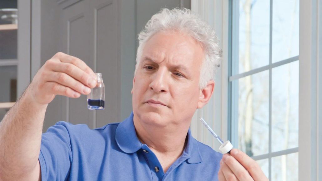 Man examining liquid in a vial, holding a dropper. He appears concerned in a well-lit indoor space.