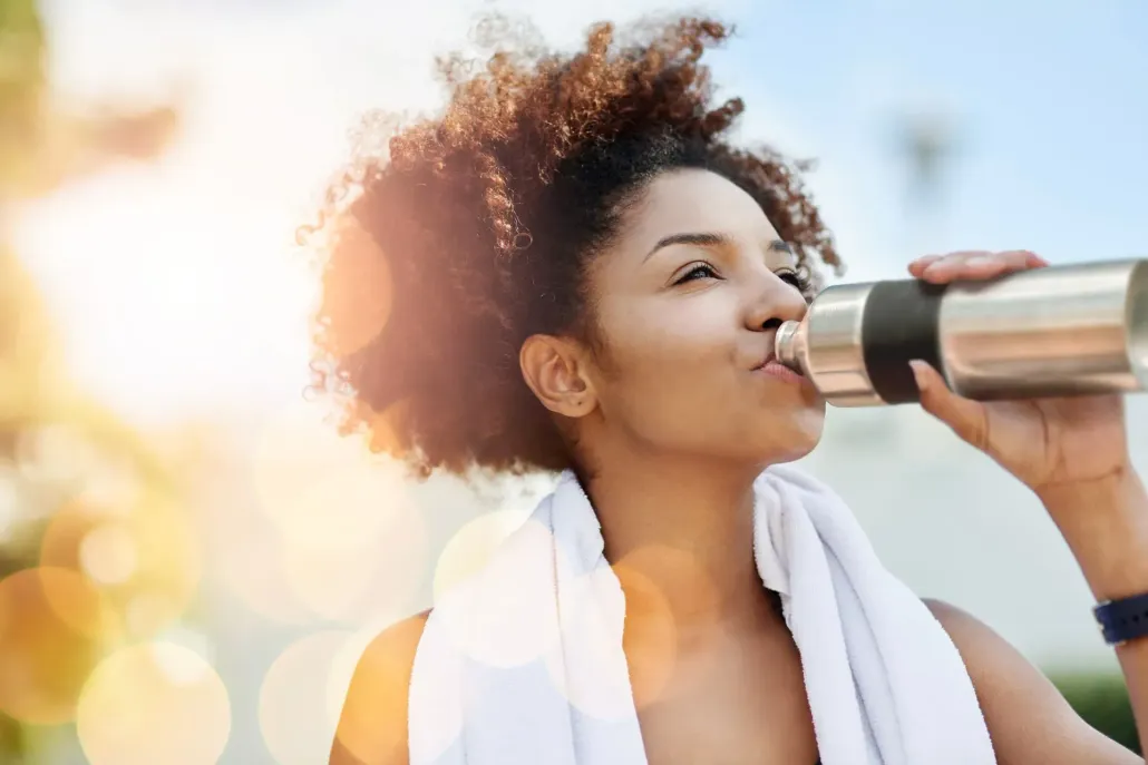 Woman drinking from a water bottle after a workout, sunny outdoor setting.