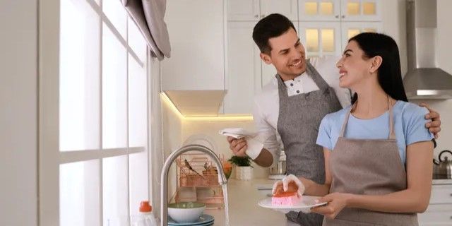 Couple washing dishes in a white kitchen, smiling. Woman washes, man embraces. Bright natural light.