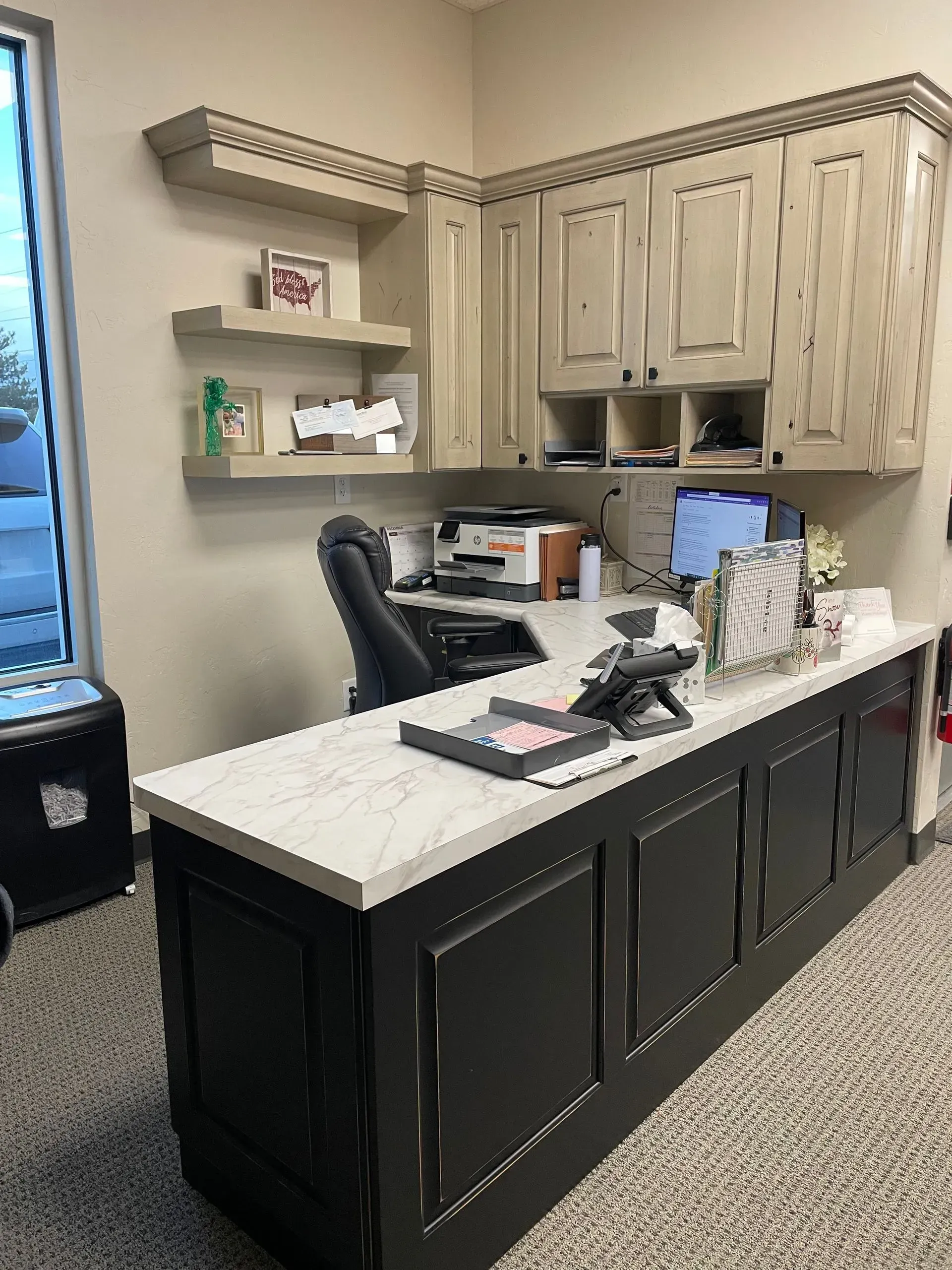 Office desk with black cabinets, white countertop, cream-colored overhead cabinets, computer monitor, and a window.