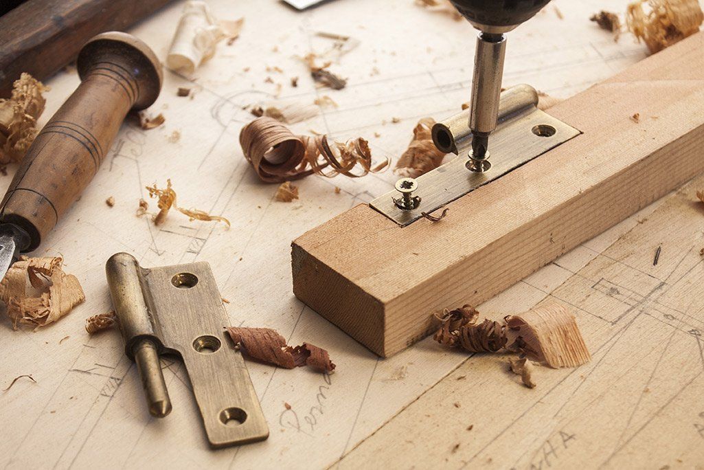 Wooden hinge being screwed into a wooden plank, surrounded by woodworking tools and shavings.