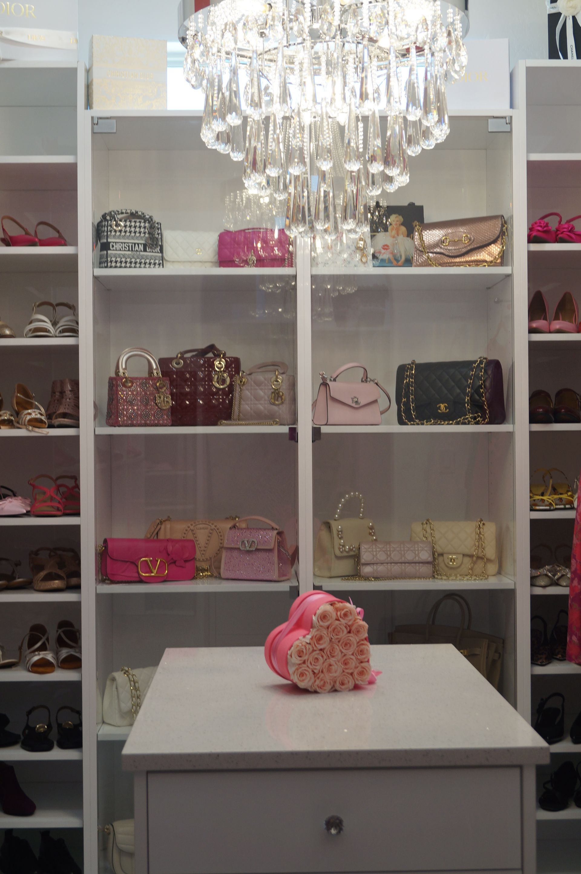Boutique display room with handbags on shelves and a crystal chandelier over a white table with pink flowers