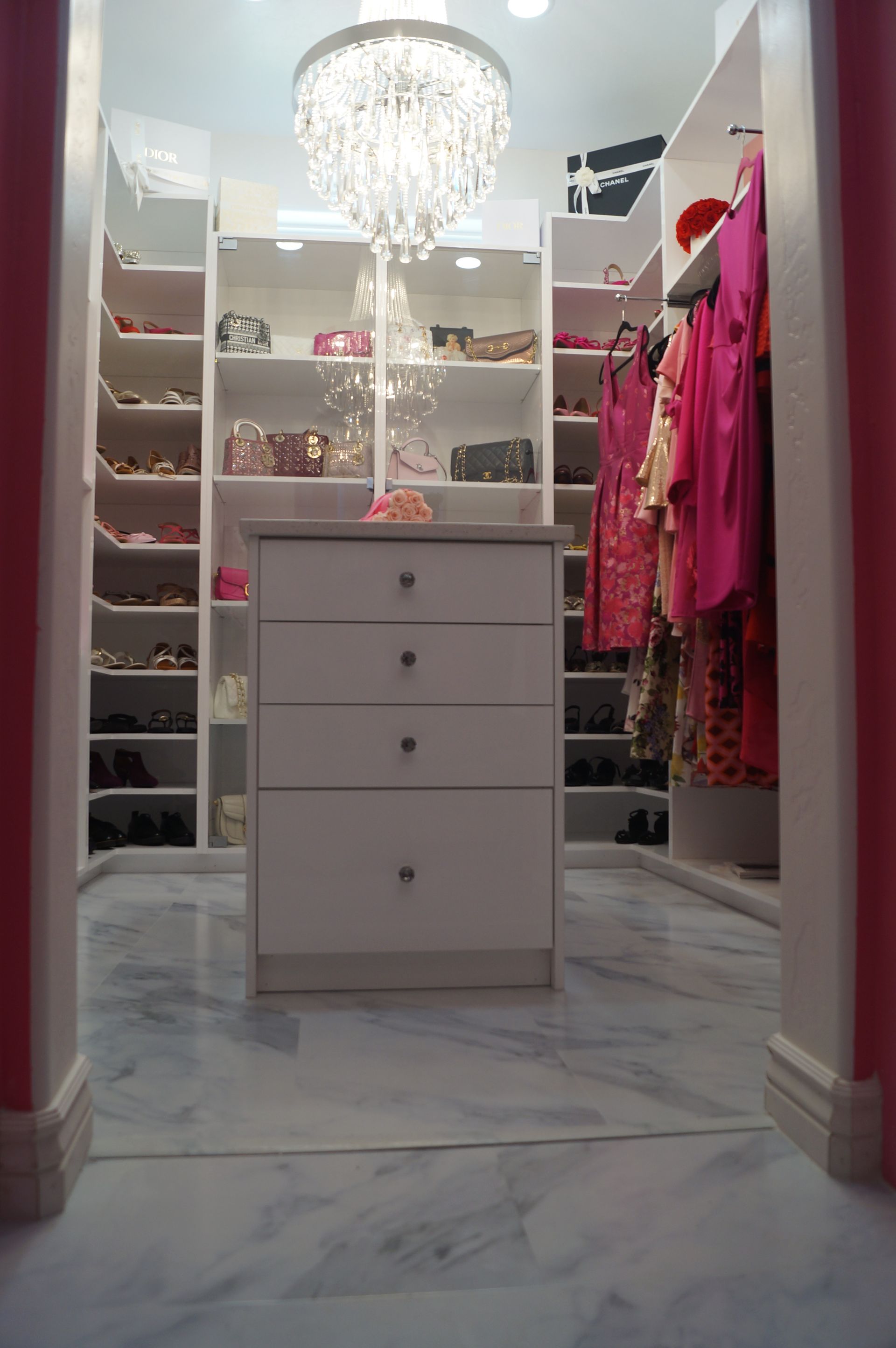 Walk-in closet with white drawers, mirrored walls, shelves of shoes, and pink clothes under a crystal chandelier