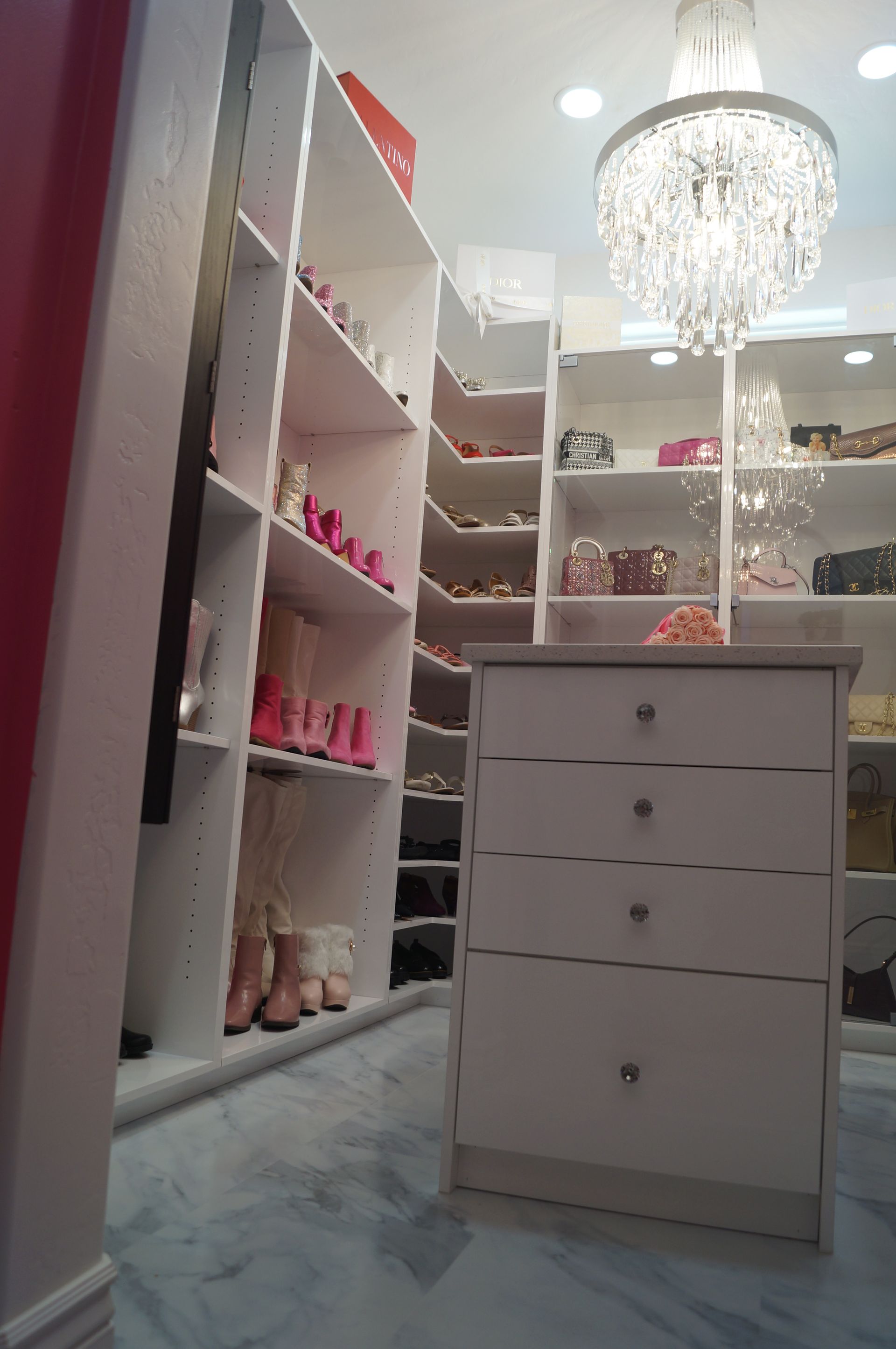 White vanity room with shelves of pink shoes, mirrored cabinets, drawers, and a chandelier