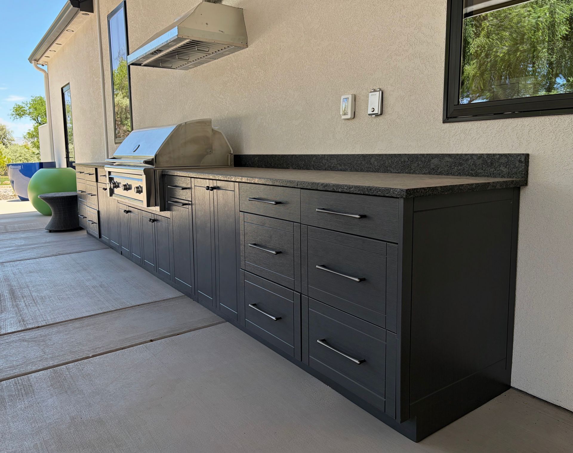Outdoor patio with a long black outdoor kitchen, grill, cabinets, and granite countertop against a beige wall